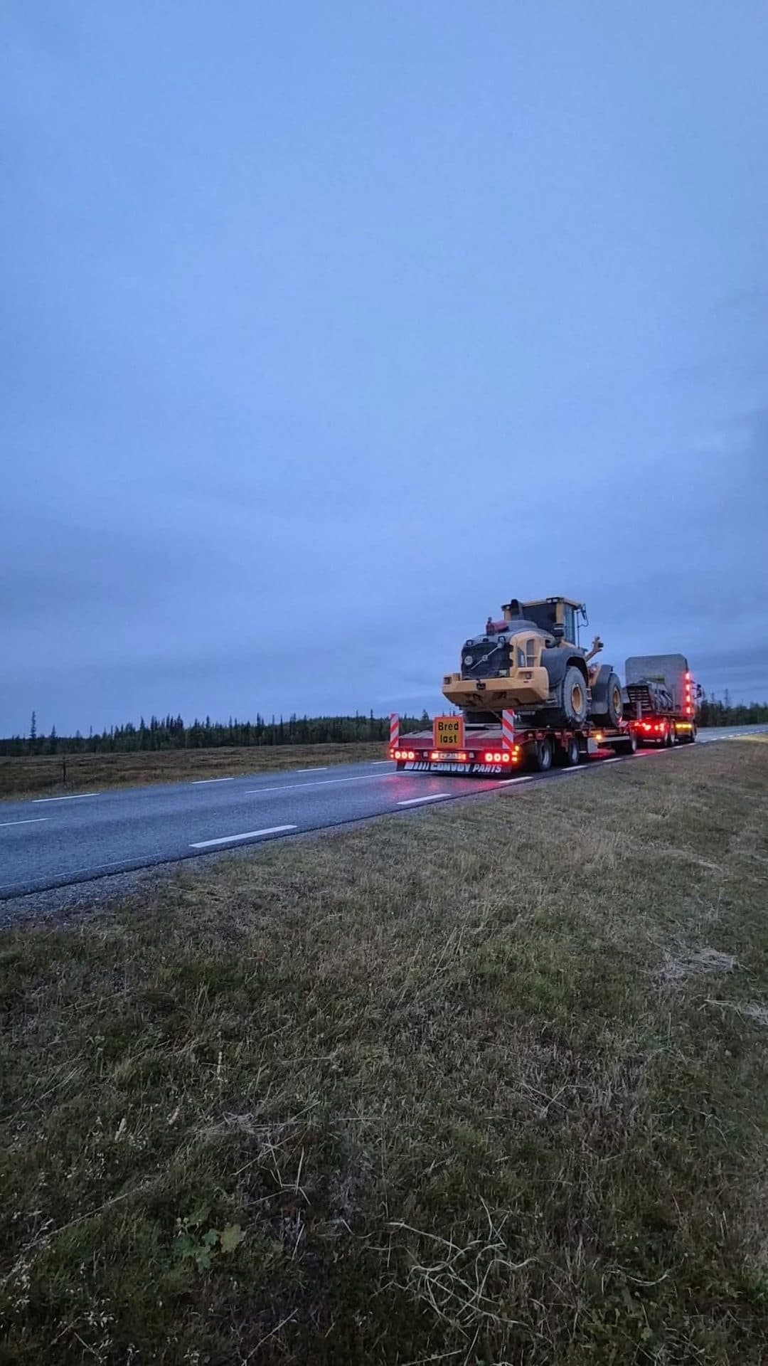 Maskinsläpet bakifrån med en hjullastare och tända ljus vid vägen i skymningen