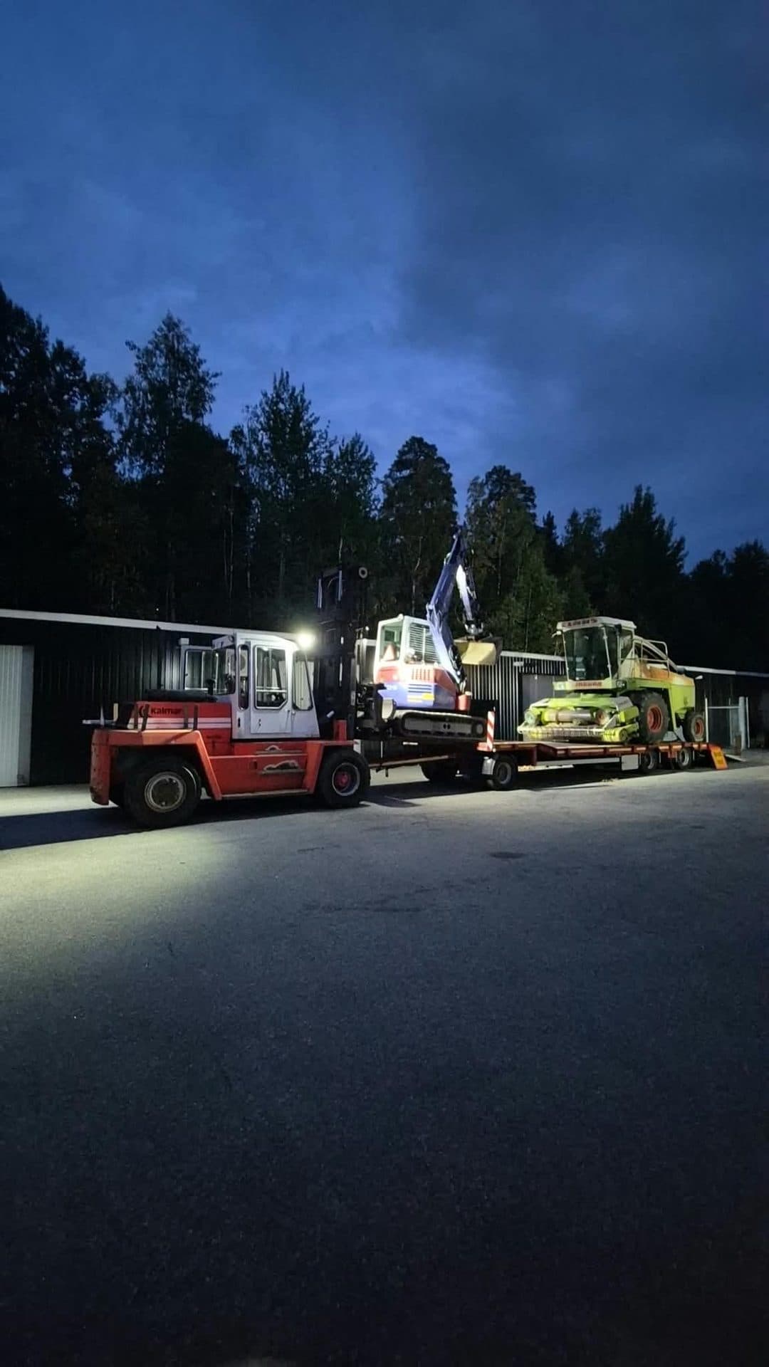 En gård i mörkret i strålkastarljus, med en truck och maskiner på maskinsläpet