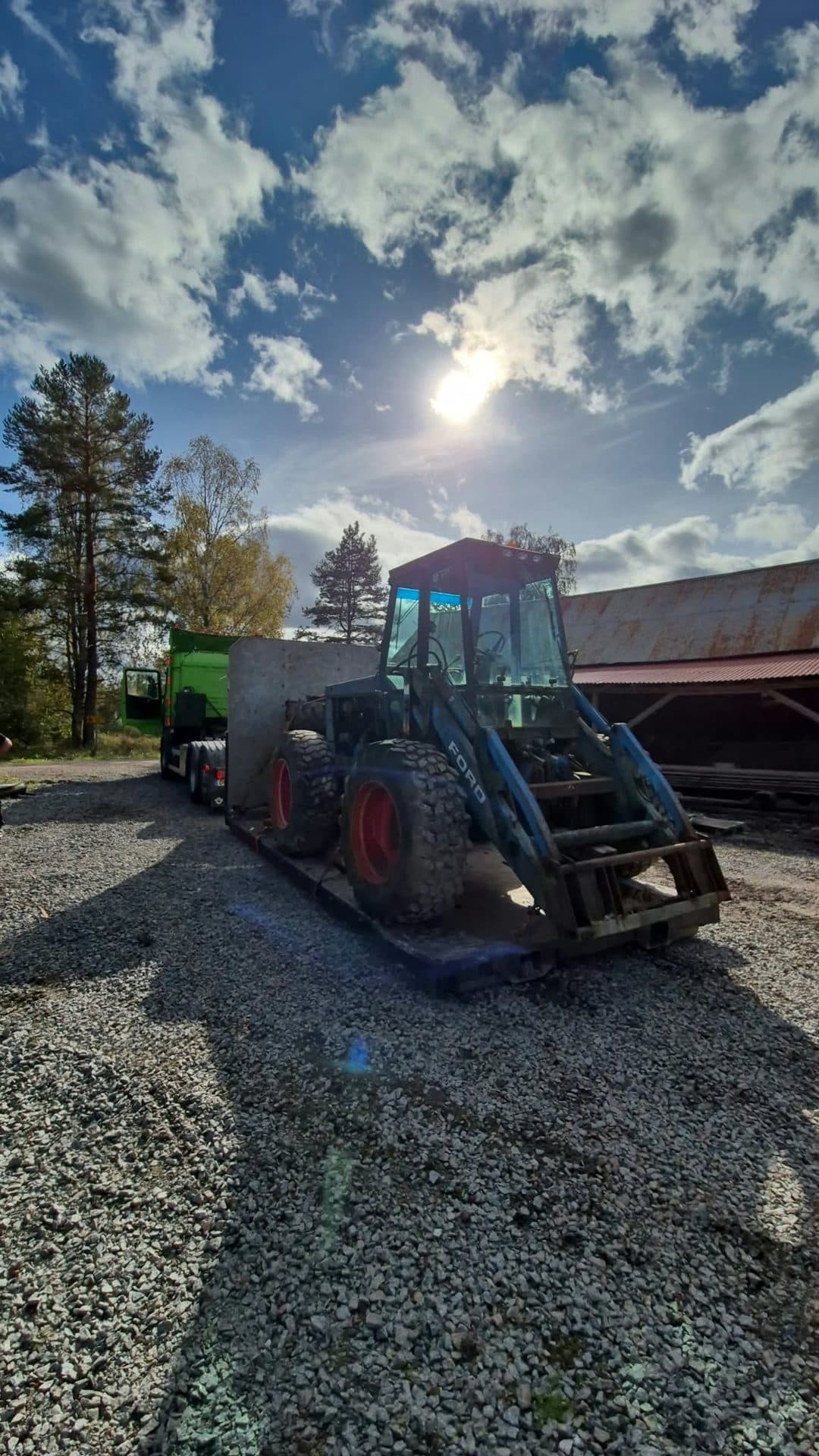 En blå traktor med frontlastare på maskinsläpet vid en röd lada, med solen bakom molnen