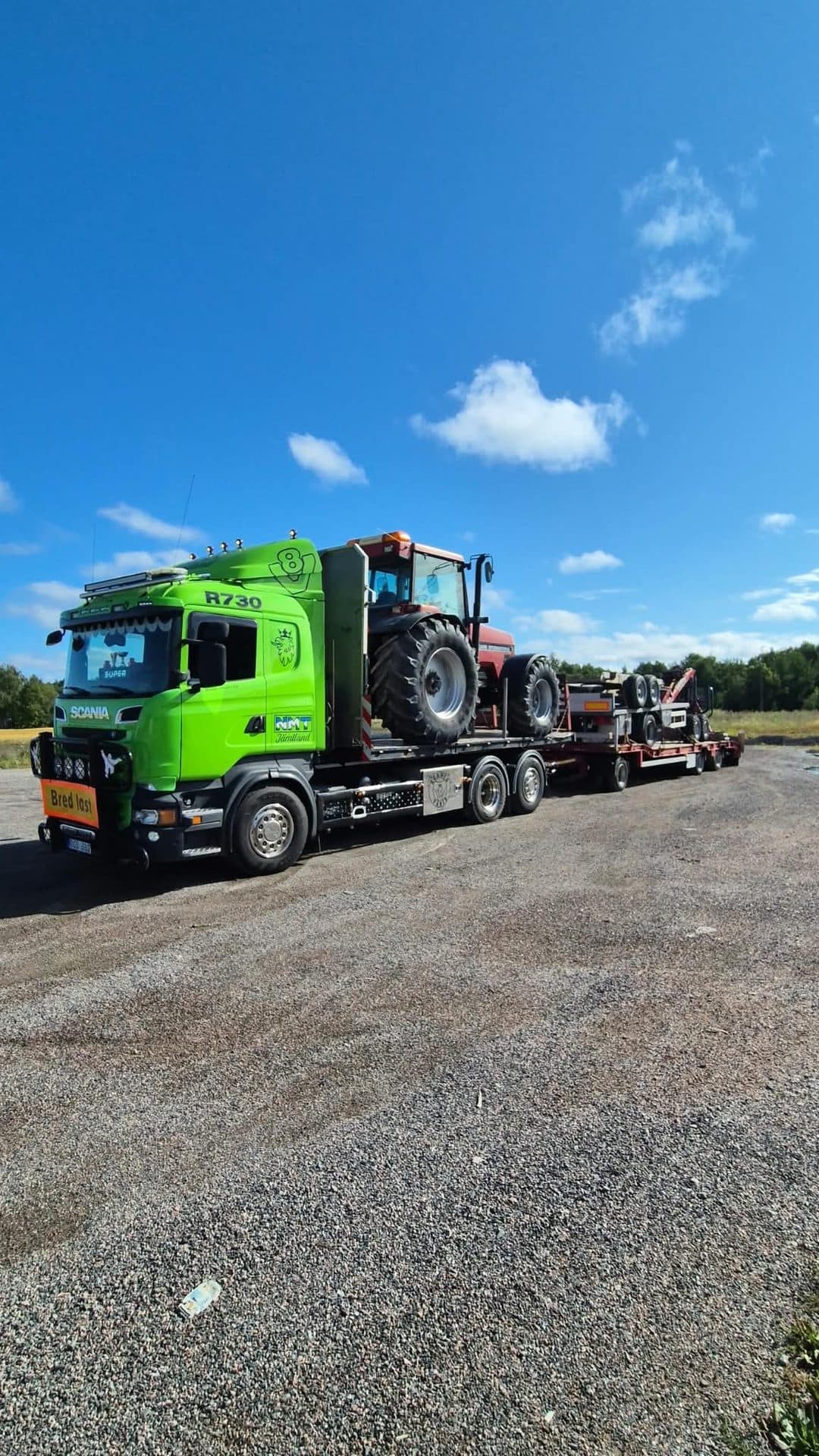 Scanian med en röd traktor på flaket och maskinsläpet bakom, skylten Bred last framtill, under blå himmel